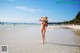 A woman in a bikini and straw hat walking on the beach.